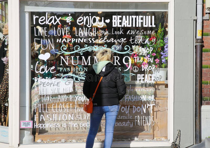 Roermond, Netherlands - February 9. 2022: Back view on one blonde caucasian woman with black winter jacket looking storefront display during window shopping on sunny day. Roermond, Netherlands - February 9. 2022: Back view on one blonde caucasian woman with black winter jacket looking storefront display during window shopping on sunny day