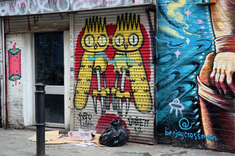 A bag of rubbish and an empty bed of cardboard where a homeless person spent the night outside a shop front covered in urban street art with a painted scene of surreal owls. Located near to Brick Lane in East London. UK. A bag of rubbish and an empty bed of cardboard where a homeless person spent the night outside a shop front covered in urban street art with a painted scene of surreal owls. Located near to Brick Lane in East London. UK