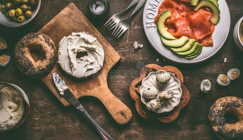 Bagel bun spread with cream-cheese on rustic breakfast table with ingredients: salmon, avocado, hummus and quail eggs stock photography