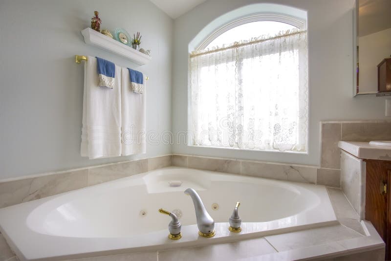 Bathtub with with gold and silver faucet beside an arched window with curtain. Ornaments on a shelf and toweld on a golden rod can be seen against the white wall. Bathtub with with gold and silver faucet beside an arched window with curtain. Ornaments on a shelf and toweld on a golden rod can be seen against the white wall.