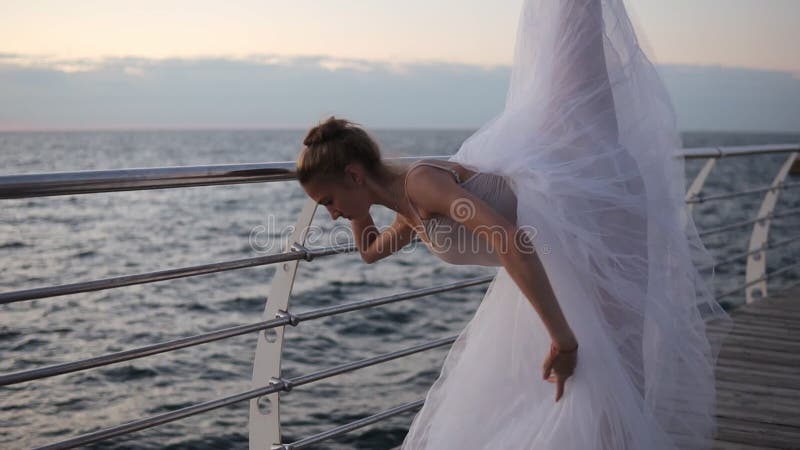 A bailarina graciosa em um tutu branco estica na frente da barra transversal perto do mar Dançarino de bailado novo bonito