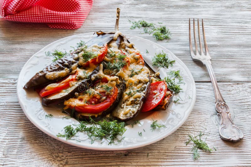 Baked eggplant in a fan shape on a white plate and vintage fork. Wooden background. Cooked with tomatoes and cheese
