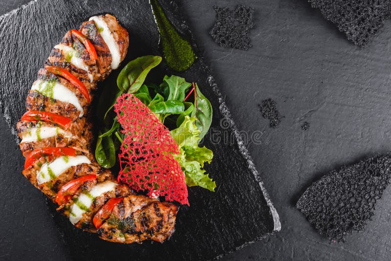Baked Veal with mozzarella cheese, tomatoes, pesto sauce and green salad on black sbackground. Hot Meat Dishes. Top view, flat lay stock images Baked Veal with mozzarella cheese, tomatoes, pesto sauce and green salad on black sbackground. Hot Meat Dishes. Top view, flat lay stock images