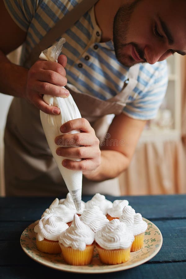 Portrait of male chef bending over kitchen table decorating freshly baked golden muffins with white cream using icing bag. Portrait of male chef bending over kitchen table decorating freshly baked golden muffins with white cream using icing bag