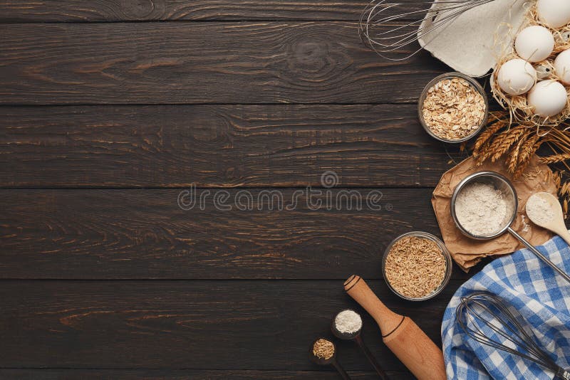 Cooking ingredients background. Border of flour and wheat grains in bowls, eggs and whisk on dark rustic wood with copy space. Dough preparing and pastry concept, top view. Cooking ingredients background. Border of flour and wheat grains in bowls, eggs and whisk on dark rustic wood with copy space. Dough preparing and pastry concept, top view