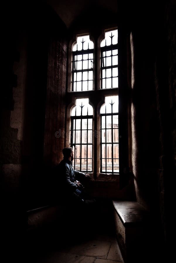 Bakre sikt på konturn av mannen som sitter i mörkret som ser till och med gammalt ljust fönster med inkommande strålar av ljus royaltyfri fotografi Bakre sikt på konturn av mannen som sitter i mörkret som ser till och med gammalt ljust fönster med inkommande strålar av ljus royaltyfri fotografi