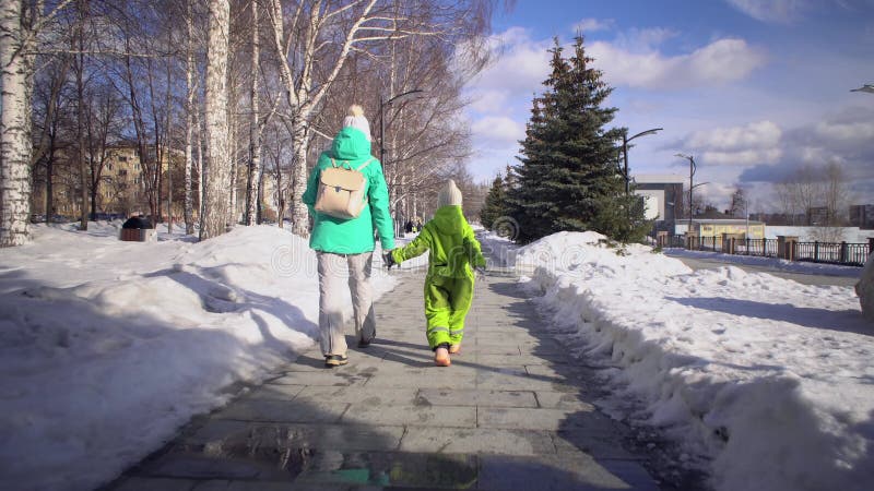 Bakåtsikt av mamma och barn går hand i hand på sidpromenaden med snöborrningar på vintern