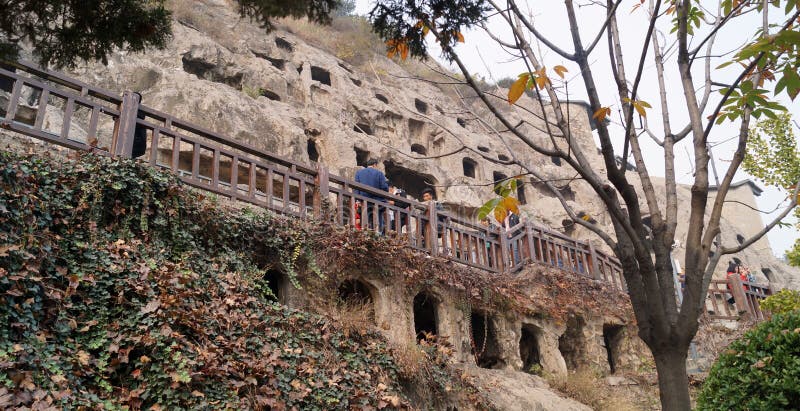 Longmen grottoes with several niches carved in the wall at the face of a mountain, buddhist temple whit several sculptures in it and a fence in front of it. Longmen grottoes with several niches carved in the wall at the face of a mountain, buddhist temple whit several sculptures in it and a fence in front of it