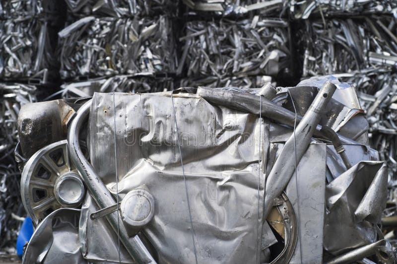 A large bale of crushed, recycled aluminum scrap viewed close-up with piled bales of scrap, crushed aluminum stack high in the background. A large bale of crushed, recycled aluminum scrap viewed close-up with piled bales of scrap, crushed aluminum stack high in the background.