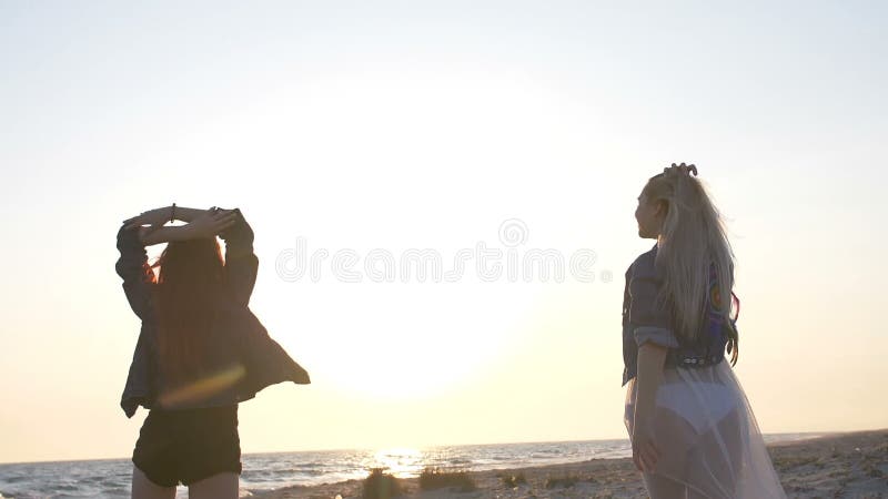 Ballo sulla spiaggia di due giovani donne sexy vestite nello stile di boho alla luce di tramonto sopra il mare