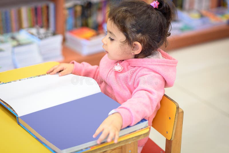 Bambina all'interno in Front Of Books Giovane bambino sveglio che si siede su una sedia vicino alla Tabella ed al libro di lettur fotografie stock Bambina all'interno in Front Of Books Giovane bambino sveglio che si siede su una sedia vicino alla Tabella ed al libro di lettur fotografie stock