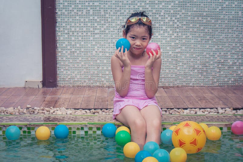 Bambina asiatica che si siede sul bordo dello stagno, sul vestito rosa d'uso dello swimminh e sulla sua mano tenenti piccola pall immagine stock Bambina asiatica che si siede sul bordo dello stagno, sul vestito rosa d'uso dello swimminh e sulla sua mano tenenti piccola pall immagine stock