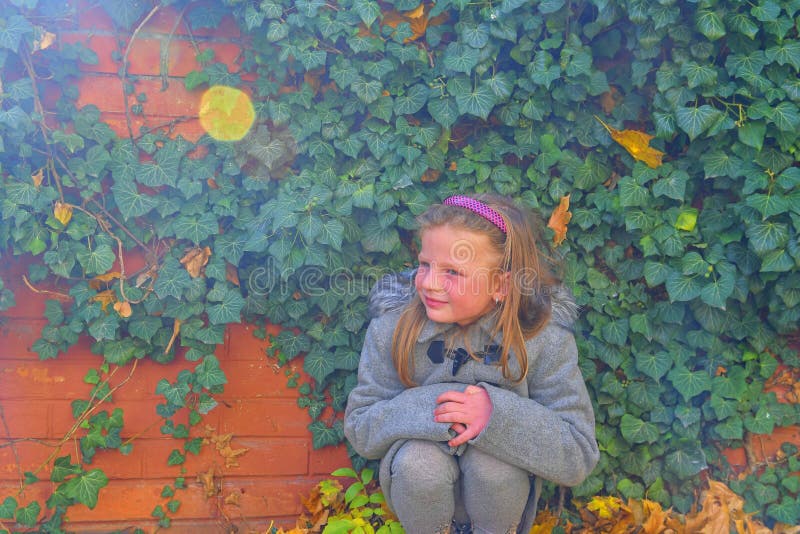 Bambina che porta retro cappotto e che si accovaccia davanti al muro di mattoni in autunno Parete dell'edera in autunno Concetto immagine stock Bambina che porta retro cappotto e che si accovaccia davanti al muro di mattoni in autunno Parete dell'edera in autunno Concetto immagine stock