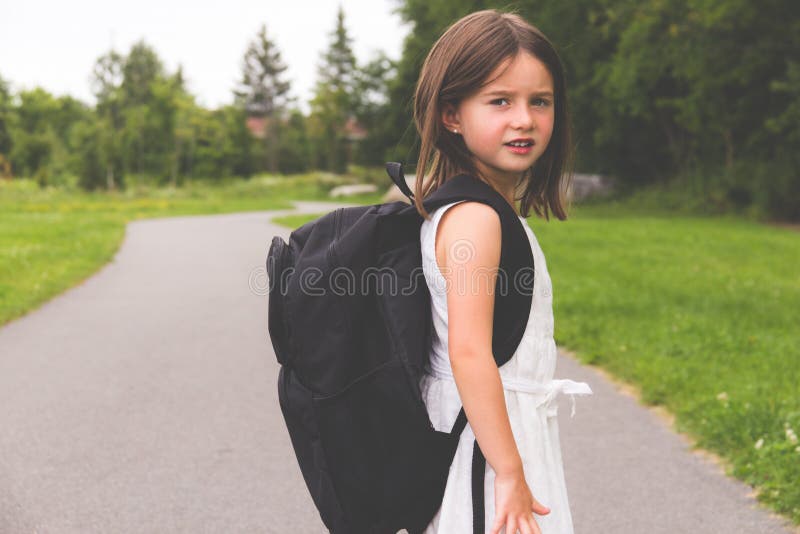 Bambina con lo zaino della scuola sul percorso che guarda di nuovo alla macchina fotografica fotografia stock Bambina con lo zaino della scuola sul percorso che guarda di nuovo alla macchina fotografica fotografia stock