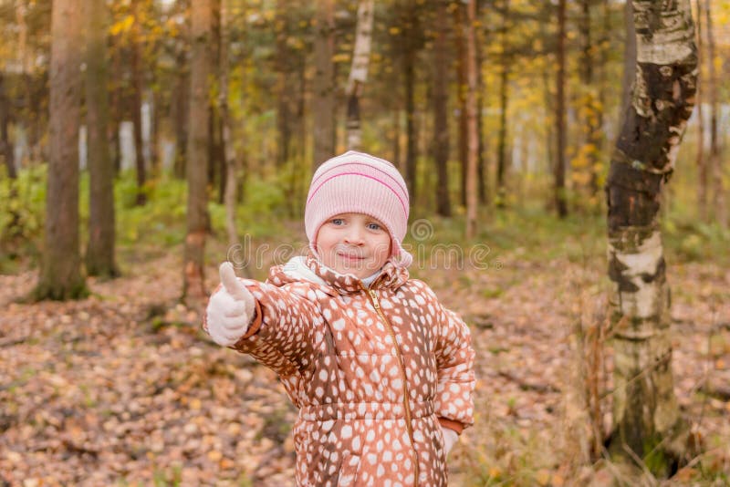 Bambina felice che mostra i pollici su nel giorno soleggiato sul fondo di autunno Alberi e lago dorati di autunno Autumn Landscap immagini stock libere da diritti Bambina felice che mostra i pollici su nel giorno soleggiato sul fondo di autunno Alberi e lago dorati di autunno Autumn Landscap immagini stock libere da diritti