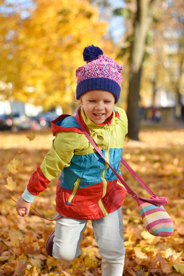 Bambina felice in vestiti luminosi che giocano con le foglie in un parco della città in autunno fotografie stock Bambina felice in vestiti luminosi che giocano con le foglie in un parco della città in autunno fotografie stock