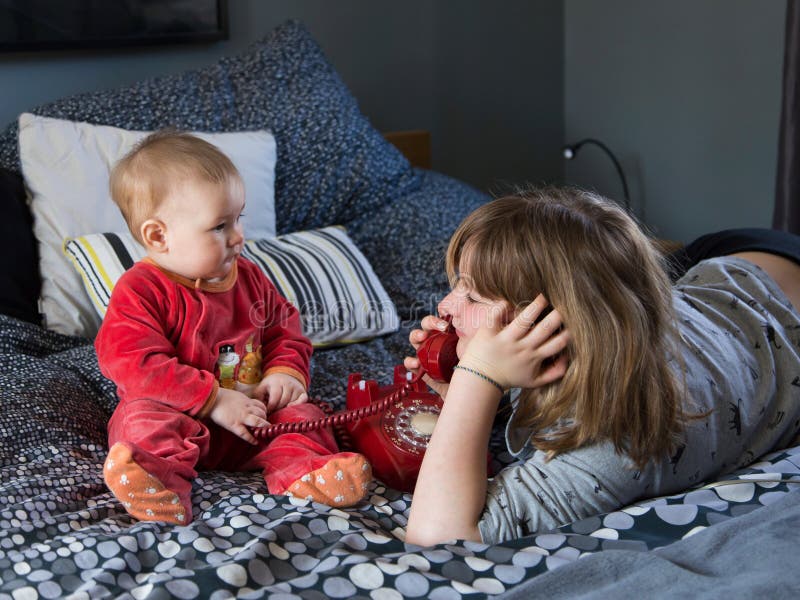 Bambina sorridente graziosa che si trova sul letto che gioca con il telefono rosso d'annata e sua sorella paffuta sveglia del bam fotografia stock libera da diritti Bambina sorridente graziosa che si trova sul letto che gioca con il telefono rosso d'annata e sua sorella paffuta sveglia del bam fotografia stock libera da diritti