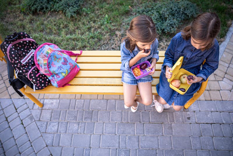 Bambine che vanno a pranzo in panchina nel cortile della scuola e mangiano dalle scatole del pranzo immagine stock