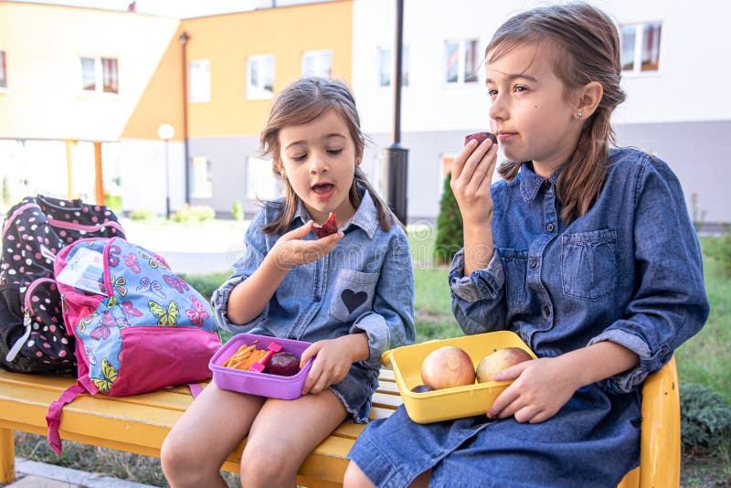 Bambine che vanno a pranzo in panchina nel cortile della scuola e mangiano dalle scatole del pranzo fotografie stock libere da diritti