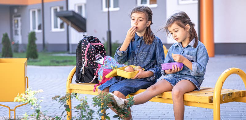 Bambine che vanno a pranzo in panchina nel cortile della scuola e mangiano dalle scatole del pranzo immagini stock libere da diritti