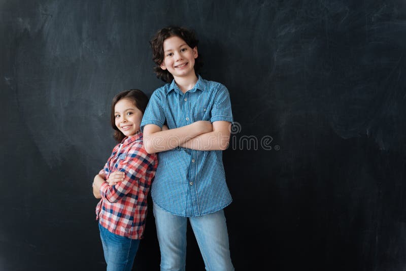 Welcome to our team. Friendly charming decisive kids standing isolated in black background and enjoying imaginary drawing while standing back to back. Welcome to our team. Friendly charming decisive kids standing isolated in black background and enjoying imaginary drawing while standing back to back