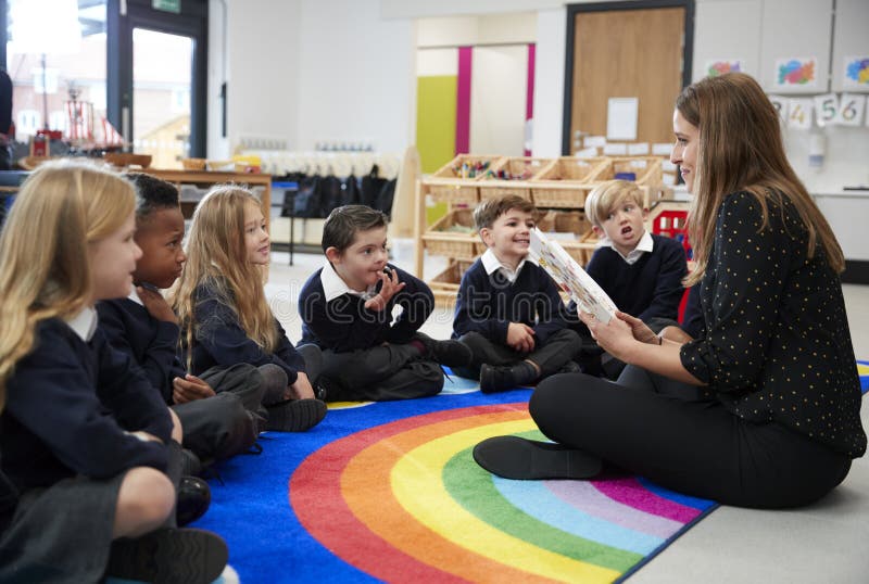 Bambini della scuola primaria che si siedono sul pavimento nella classe che ascolta il loro insegnante femminile che legge un lib immagini stock Bambini della scuola primaria che si siedono sul pavimento nella classe che ascolta il loro insegnante femminile che legge un lib immagini stock