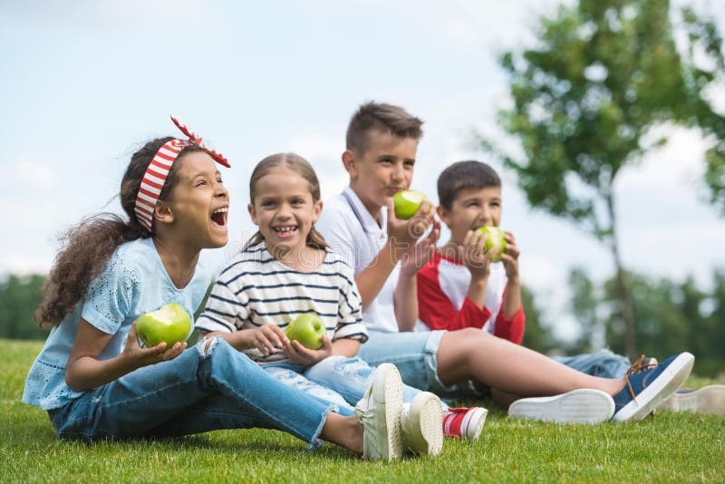 Happy multiethnic children eating green apples while sitting together on green grass. Happy multiethnic children eating green apples while sitting together on green grass