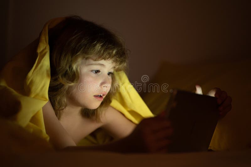 Bambino carino sdraiato sotto la coperta con una pastiglia. bambino sorpreso a letto sotto coperta mentre guarda il video su una t fotografie stock