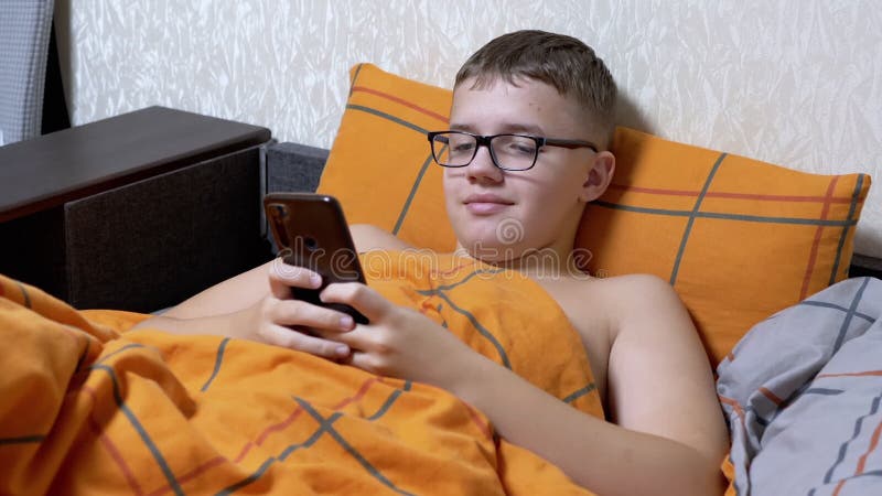 Bambino che guarda un video interessante su uno smartphone disteso in un letto accogliente