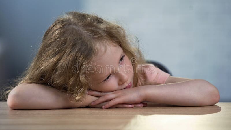 Bambino femminile riccio infelice che si siede da solo alla tavola, servizio sociale per i bambini immagini stock Bambino femminile riccio infelice che si siede da solo alla tavola, servizio sociale per i bambini immagini stock