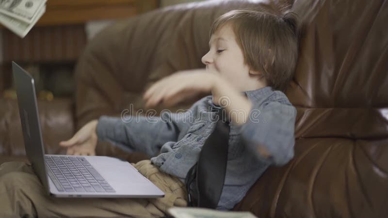 Bambino piccolo adorabile del ritratto che si siede sul sofà di cuoio con il computer portatile sulle gambe e sui soldi di lancio