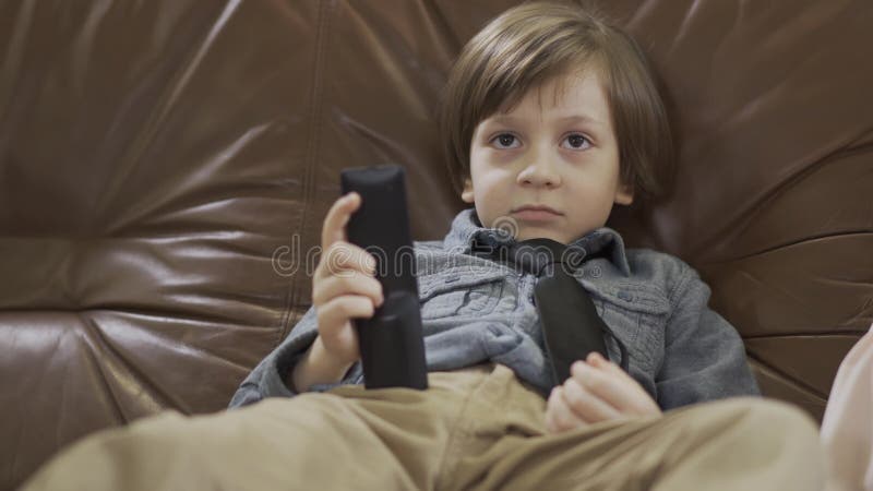 Bambino piccolo adorabile del ritratto che si siede sul sofà di cuoio con le gambe a parte che cambiano i canali sulla TV facendo