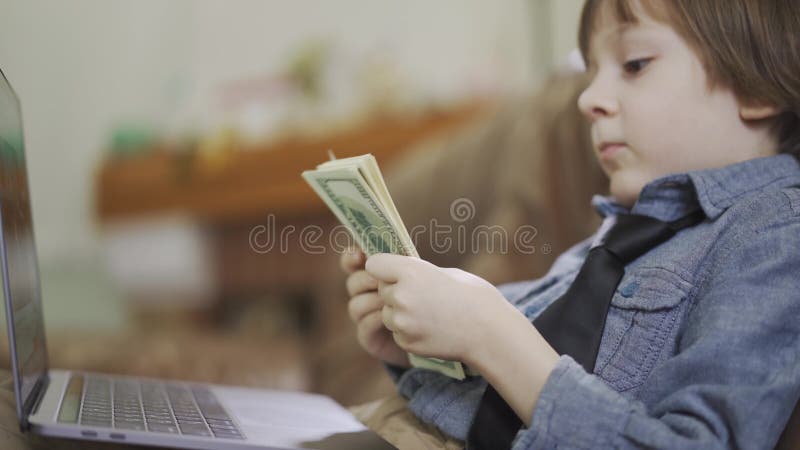 Bambino piccolo adorabile del ritratto in rivestimento dei jeans che si siede sul sofà davanti al computer portatile che conta i