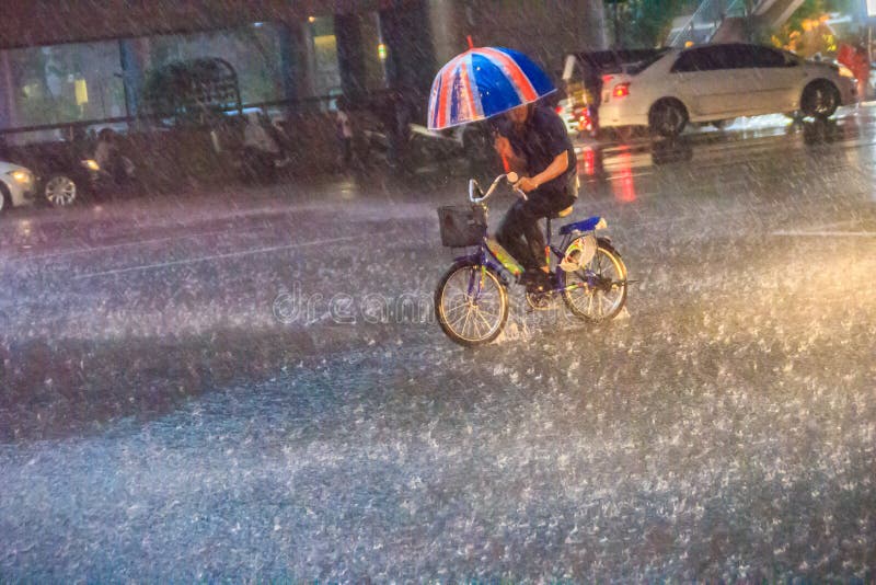 Bangkok, Thailand - July 6. 2017: A man is ridding the bicycle under a heavy rain on the street in the evening. Unidentified man is riding a bicycle and carrying an umbrella in heavy rain on a street.