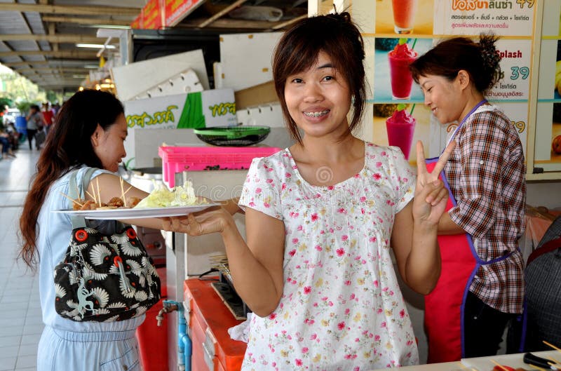 Smiling woman with braces on her teeth offers a plate of free sample pork sausages at her booth in the expansive Chatuchak Park market hall in Bangkok, Thailand. Smiling woman with braces on her teeth offers a plate of free sample pork sausages at her booth in the expansive Chatuchak Park market hall in Bangkok, Thailand.