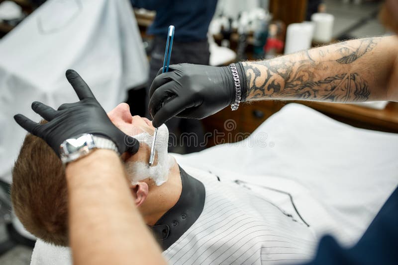 Barba tradicional. foto cortada do barbeiro em luvas pretas com um cliente de barbear de braço tatuado com uma reta perigosa imagens de stock royalty free