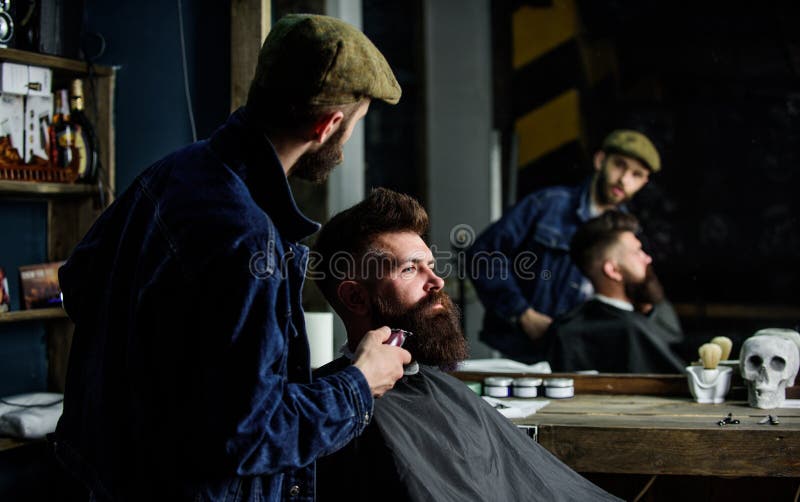 Barbeiro ocupado a cuidar da barba de um cliente hipster, reflexo no espelho ao fundo. Conceito de higiene pessoal. Hipster com ba imagem de stock