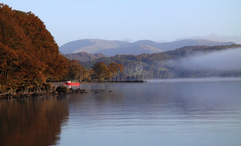 Red boat moored by the edge of Lake Coniston, Cumbria, England in the early morning mist with views of the Lake District fells beyond. Red boat moored by the edge of Lake Coniston, Cumbria, England in the early morning mist with views of the Lake District fells beyond.