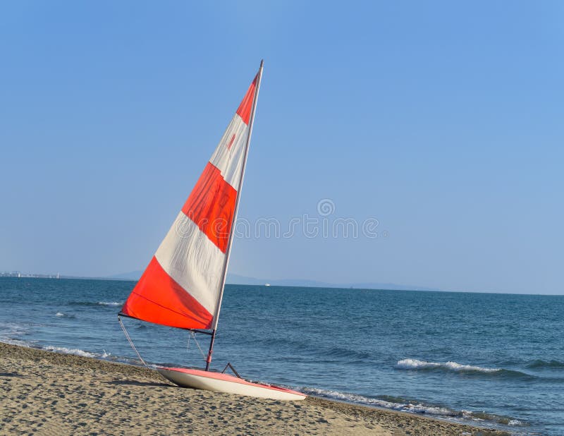 Barca a vela con la vela variopinta rossa e bianca sulla spiaggia fotografia stock libera da diritti Barca a vela con la vela variopinta rossa e bianca sulla spiaggia fotografia stock libera da diritti