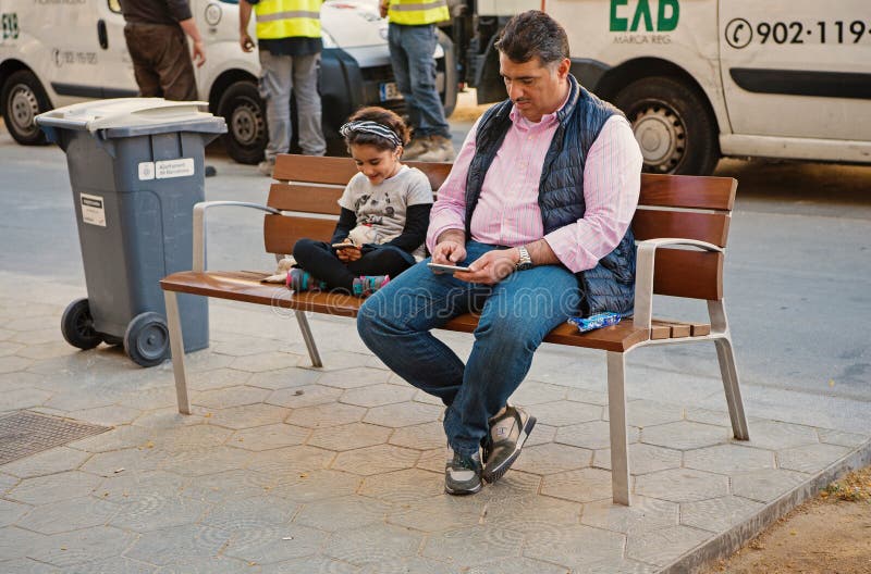 Barcelona, Espa?a - 30 de marzo de 2016: El hombre y la ni?a del padre buscan en los smartphones que se sientan en banco en la ca imágenes de archivo libres de regalías Barcelona, Espa?a - 30 de marzo de 2016: El hombre y la ni?a del padre buscan en los smartphones que se sientan en banco en la ca imágenes de archivo libres de regalías