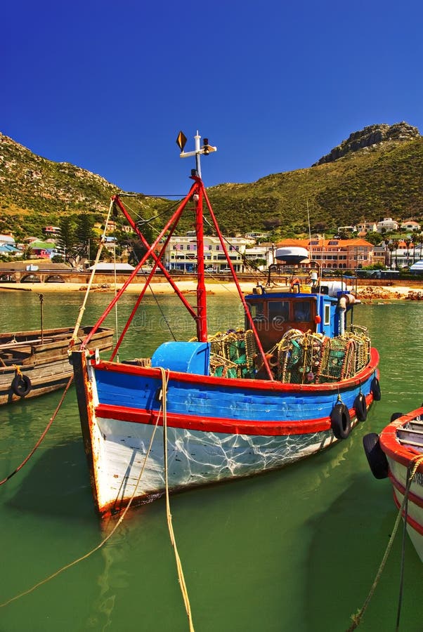 A colorful painted fishing boat swimming in the water and docking at the harbor in False Bay, South Africa, in front of little houses close to the sea. A colorful painted fishing boat swimming in the water and docking at the harbor in False Bay, South Africa, in front of little houses close to the sea