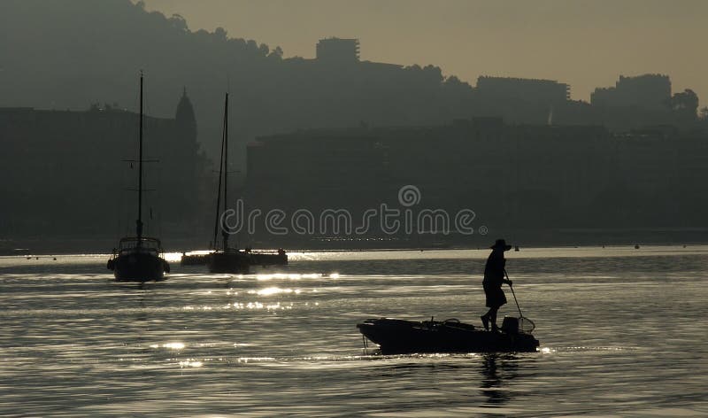 Silhouetted skyline of Cannes city at dusk with person on boat in foreground cleaning debris from sea. Silhouetted skyline of Cannes city at dusk with person on boat in foreground cleaning debris from sea.