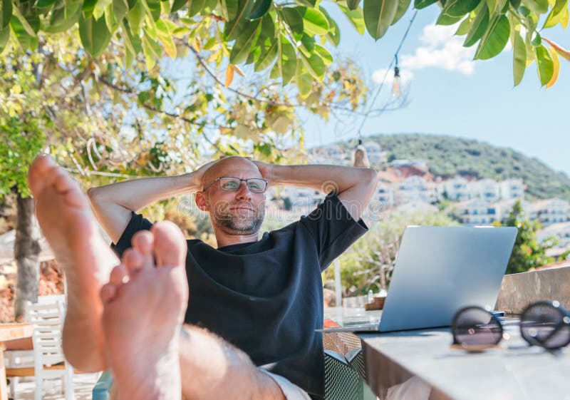 Barefoot Freelancer man enjoying remote work on sunny terrace surrounded by greenery. Digital nomad leaning back with laptop stock photography