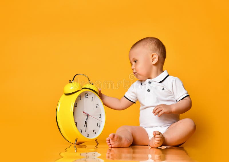 Infant baby boy toddler is occupied with a big yellow alarm clock playing looking exploring on yellow background