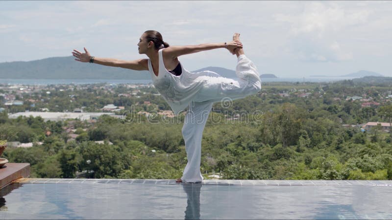 Barefooted woman in standing bow pose near swimming pool against green forested islands