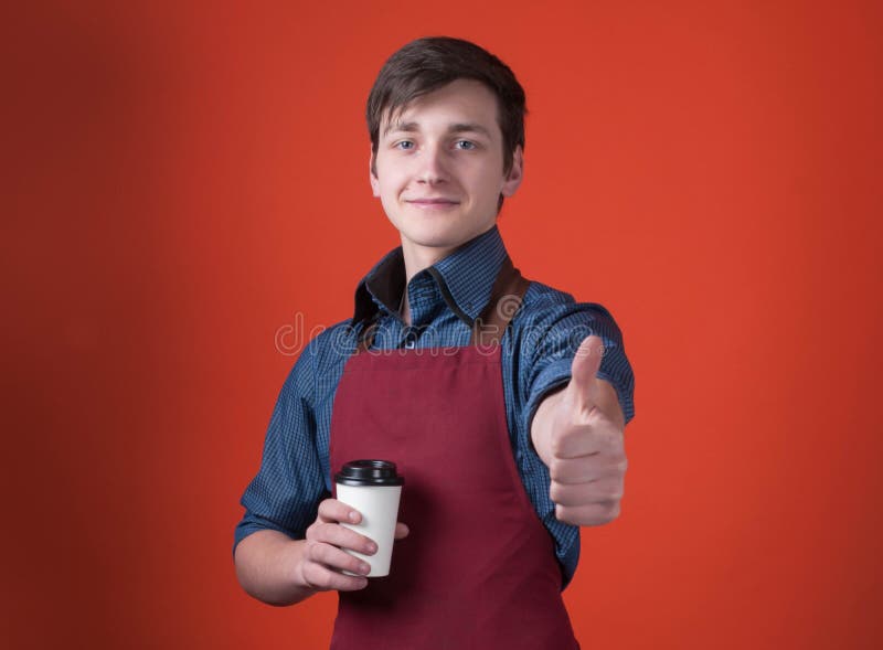 Barista bello con capelli scuri nel grembiule di Borgogna che tiene caffè in tazza di carta, esaminante macchina fotografica e sf immagine stock Barista bello con capelli scuri nel grembiule di Borgogna che tiene caffè in tazza di carta, esaminante macchina fotografica e sf immagine stock
