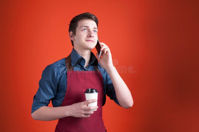 Barista med mörkt hår i det burgundy förklädet som rymmer den pappers- koppen med kaffe och talar på smartphonen arkivbilder Barista med mörkt hår i det burgundy förklädet som rymmer den pappers- koppen med kaffe och talar på smartphonen arkivbilder