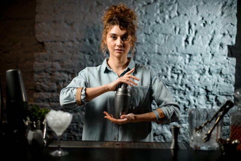 Barista professionista tiene in mano un agitatore di metallo al bar. Giovane barista professionista, camicetta blu leggera, stringe in mano un frullatore di metallo al bancone del bar
