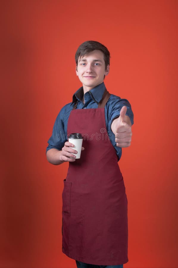 Barista χαμόγελου με τη σκοτεινή τρίχα στον κόκκινο καφέ εκμετάλλευσης ποδιών στο φλυτζάνι εγγράφου, που εξετάζει τη κάμερα και π στοκ εικόνες Barista χαμόγελου με τη σκοτεινή τρίχα στον κόκκινο καφέ εκμετάλλευσης ποδιών στο φλυτζάνι εγγράφου, που εξετάζει τη κάμερα και π στοκ εικόνες