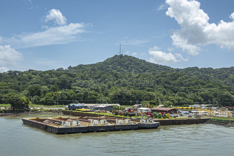 Panama Canal, Panama - July 24, 2023: Parked barges along quay with colorful collection of buoys on shore halfway canal, under blue cloudscape. Green forested hill with antenna in back. Panama Canal, Panama - July 24, 2023: Parked barges along quay with colorful collection of buoys on shore halfway canal, under blue cloudscape. Green forested hill with antenna in back